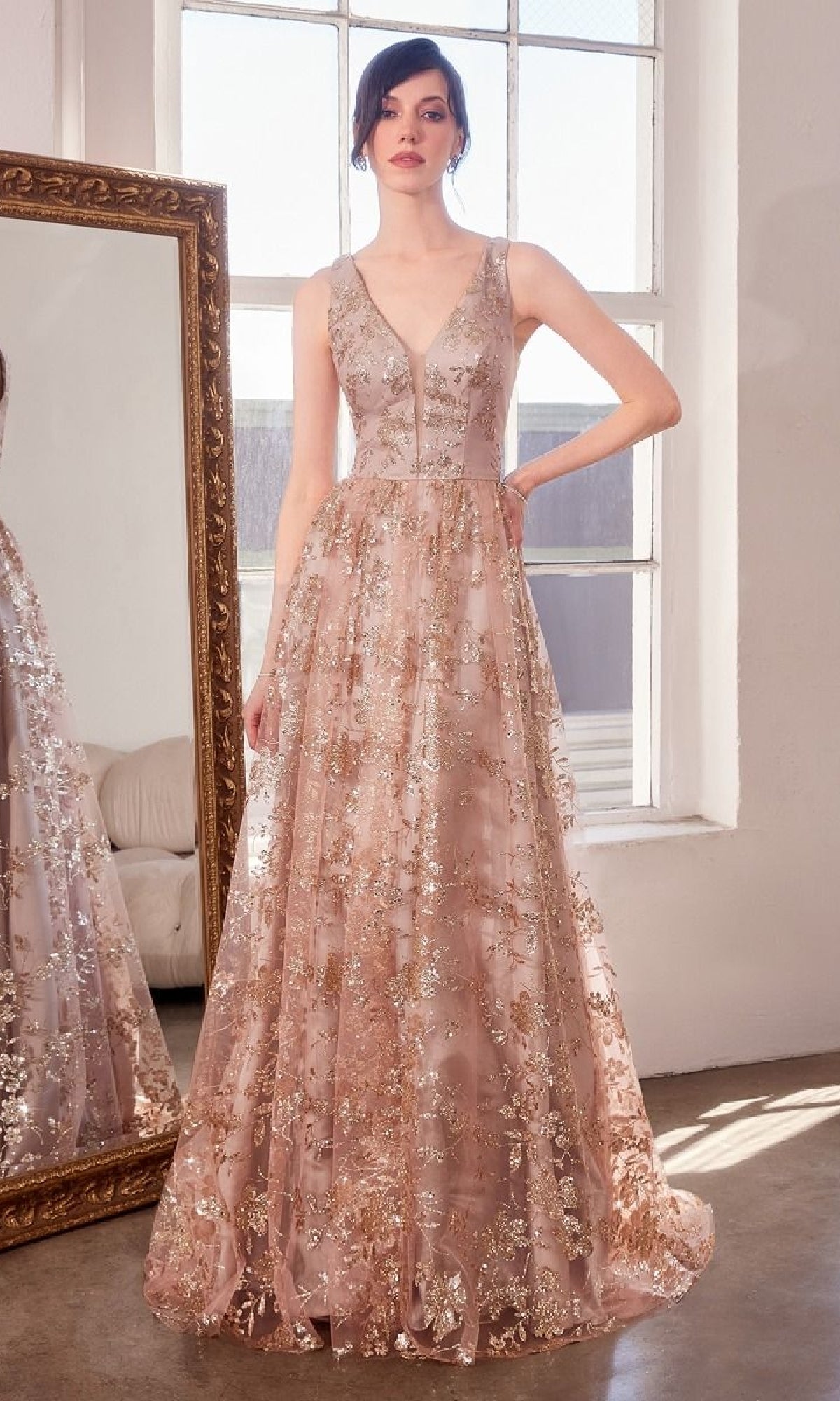 A woman stands indoors beside a large mirror and window, wearing the Embroidered-Lace Long Prom Dress: Amoris T606, a sleeveless formal gown with gold floral embroidery.